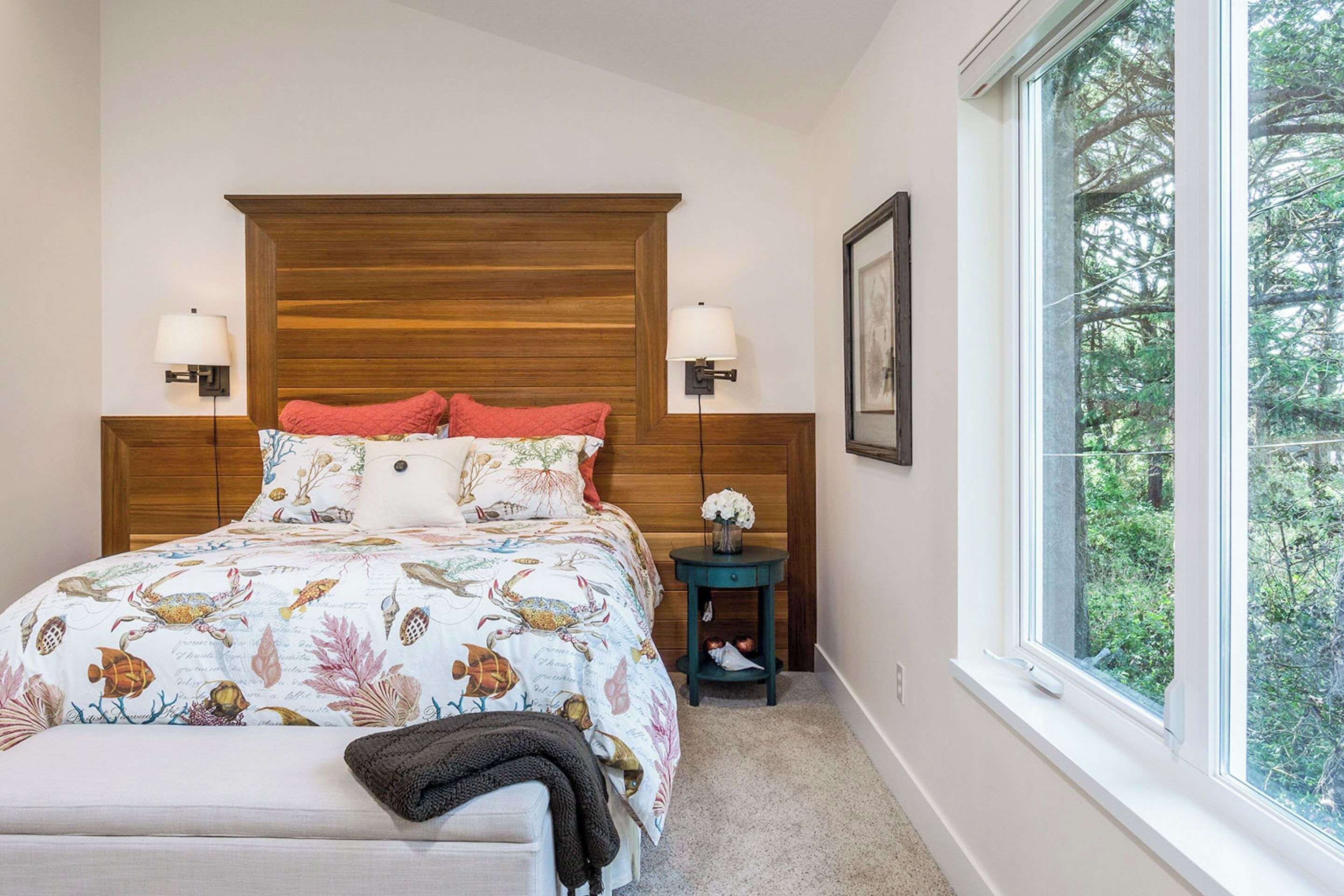 Ein Schlafzimmer mit einem Holz-Kopfteil, einem Bett mit floralem Überwurf, zwei Nachttischlampen, einem Nachttisch mit Blumenvase und einem Fenster mit Blick auf Bäume.
