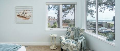 A bedroom with a bed, a chair, a window with a view of the sea, and a wall art piece.