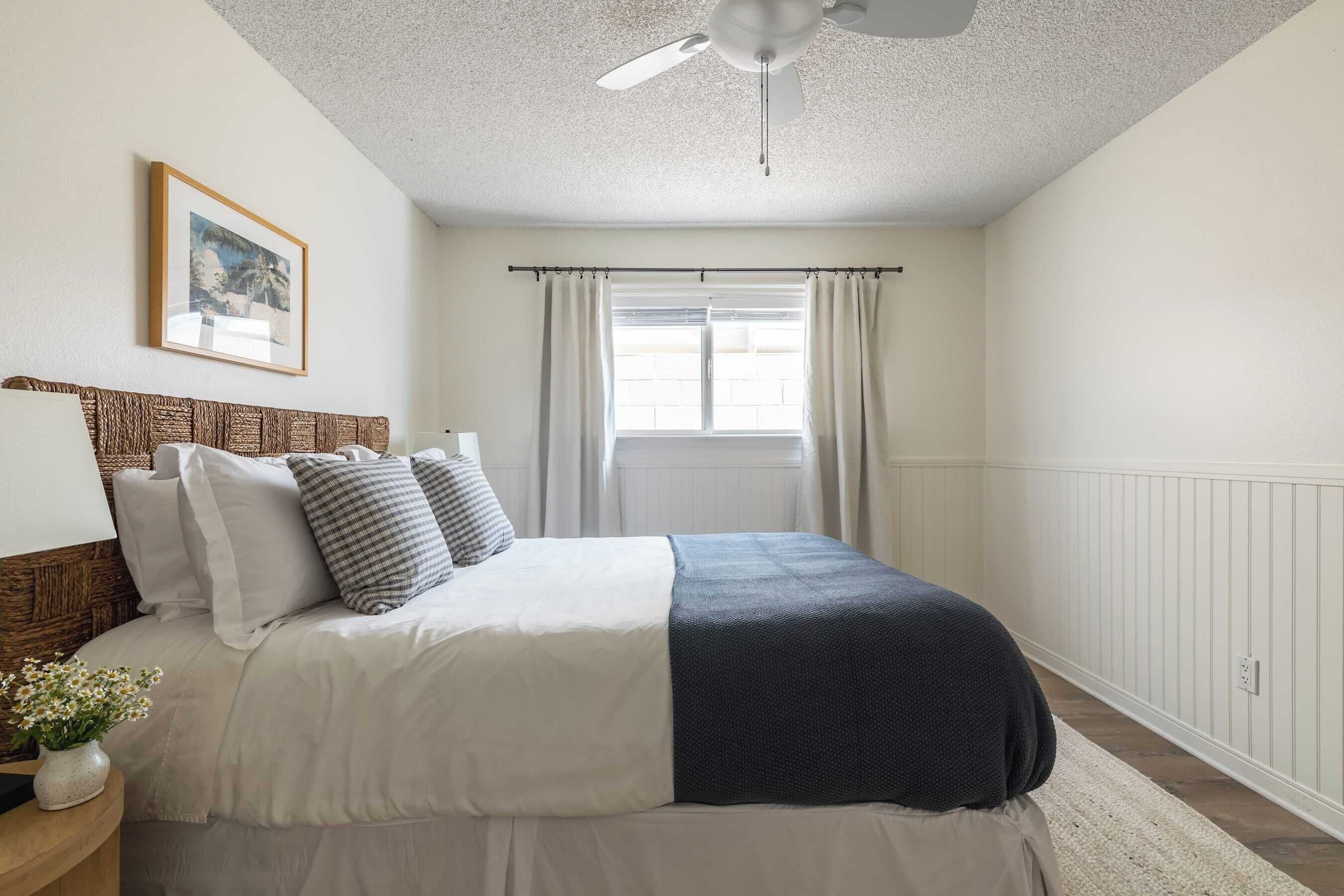 A bedroom with a bed, a ceiling fan, a window with curtains, and a framed picture on the wall.