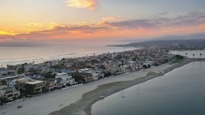 An aerial view of a coastal town with buildings along the beachfront, a pier extending into the water, and a clear sky.