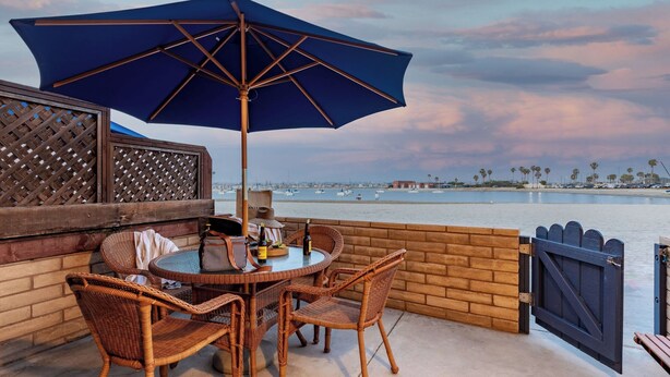 A patio with wicker furniture, a table, and a blue umbrella overlooking a waterfront with boats and palm trees. - Bay View II by Avantstay Ocean View Mission Beach Home on the Sand! (San Diego)