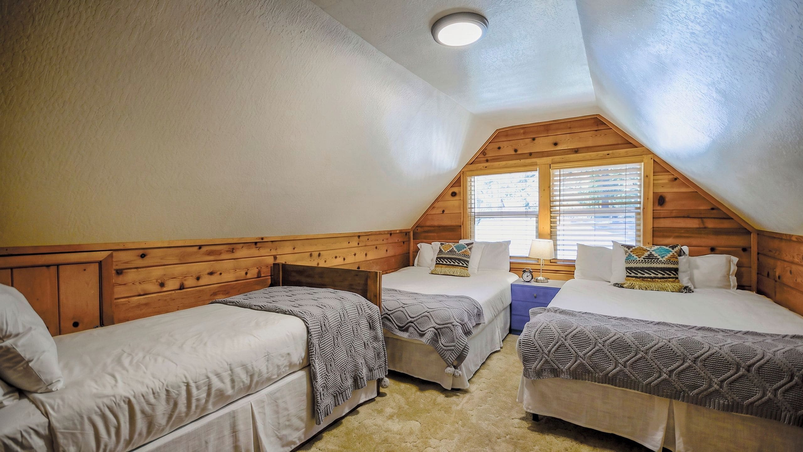 Ein Schlafzimmer mit zwei Betten, Holz-Kopfteilen, einem Fenster mit Jalousien und einer Deckenleuchte.