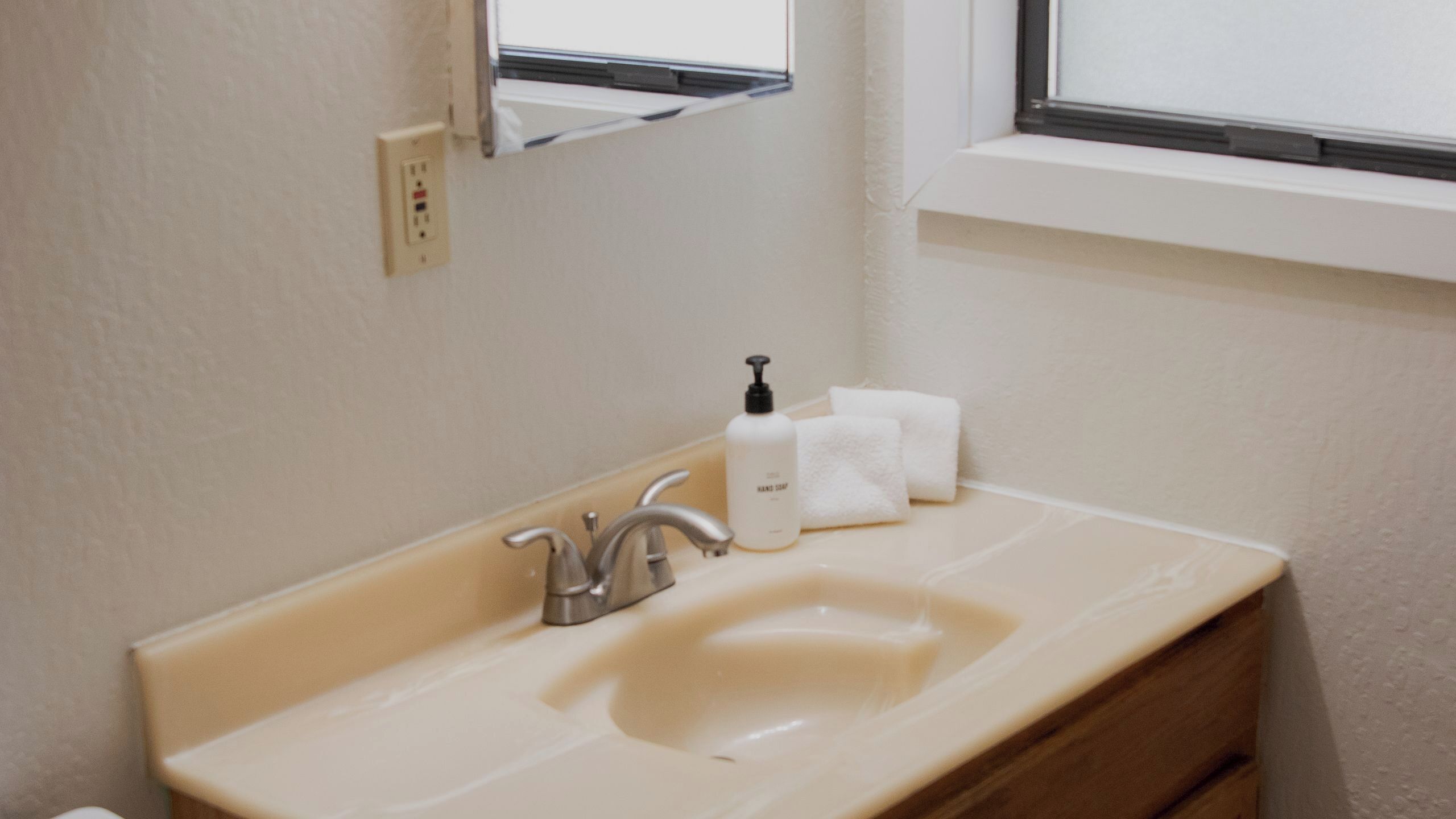 A bathroom with a beige sink, a mirror, a window, and a bottle of hand soap.