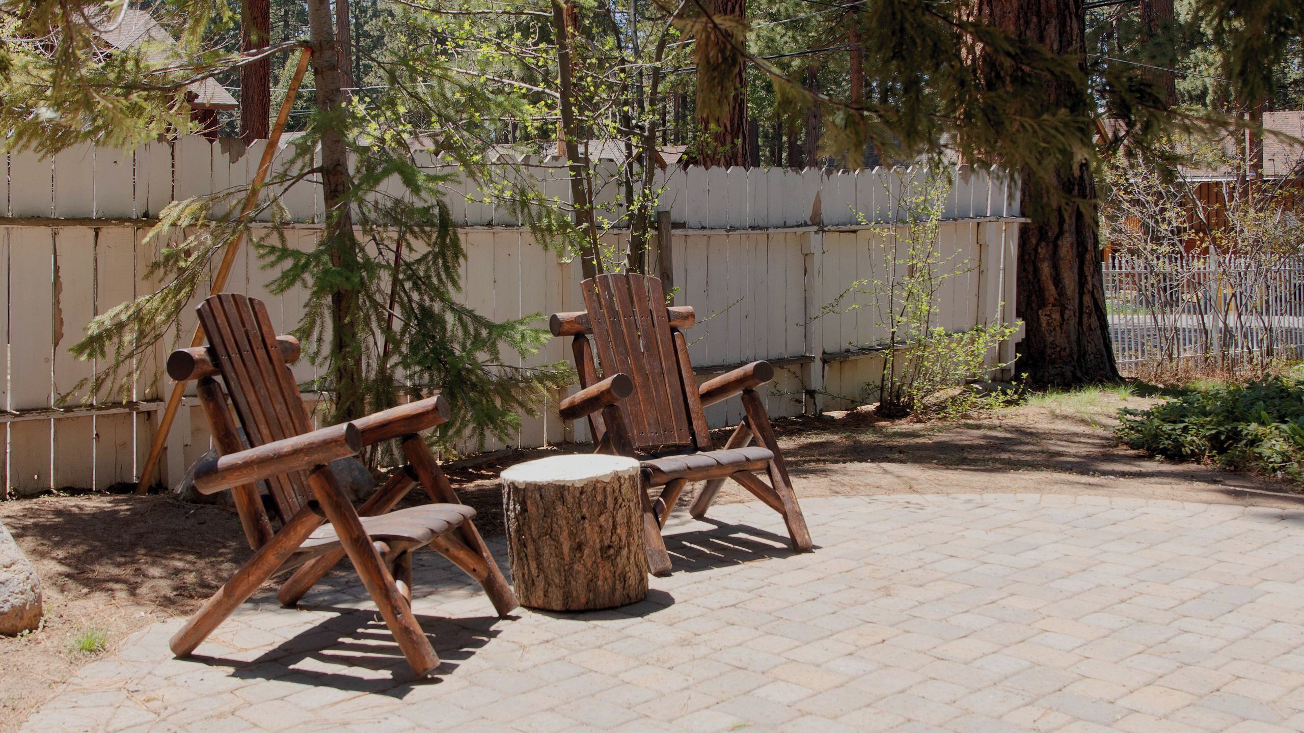 Dos sillas de madera tipo Adirondack con una mesa redonda de madera, ubicadas en un patio empedrado con una cerca blanca y árboles al fondo.