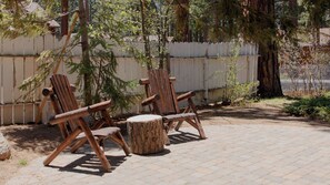 Two wooden Adirondack chairs and a round wooden table in an outdoor patio area with a white fence and trees in the background.