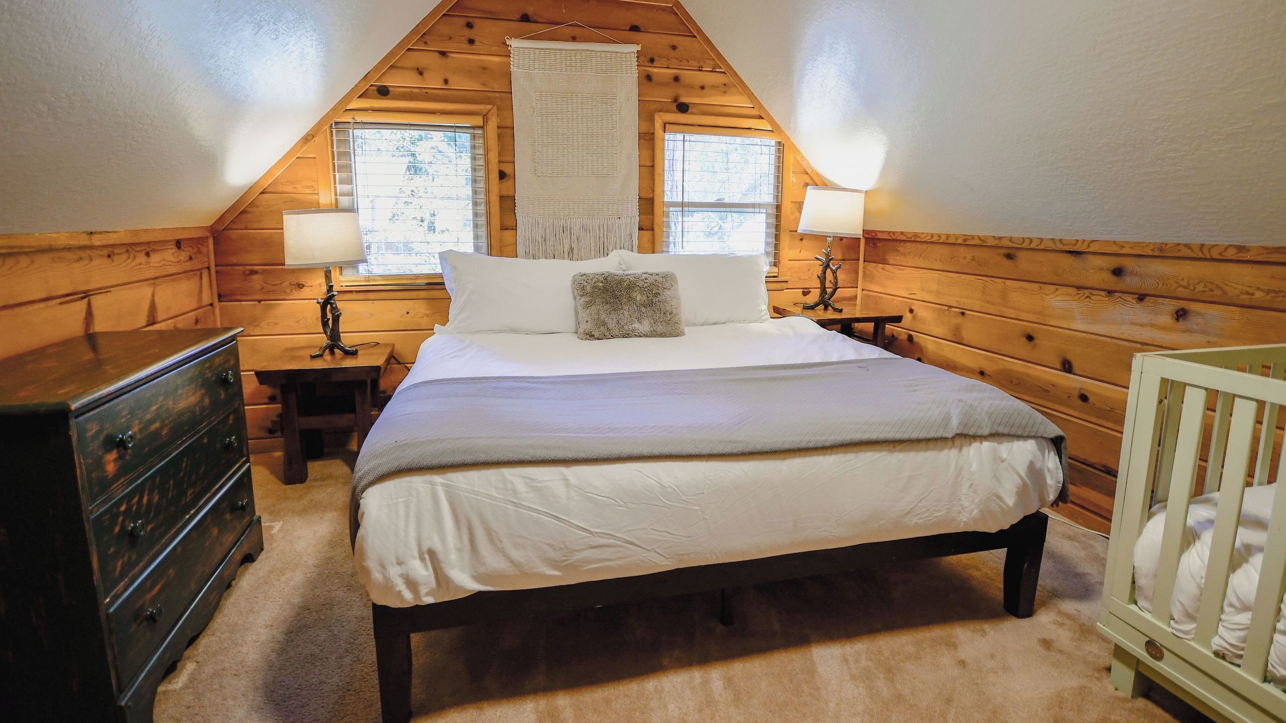 A bedroom with a wooden sloped ceiling, a bed with white bedding, two bedside lamps, a wooden dresser, and a crib.