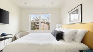 Une chambre d’hôtel avec un lit, un bureau, une chaise, une fenêtre donnant sur des immeubles et un tableau encadré au mur.