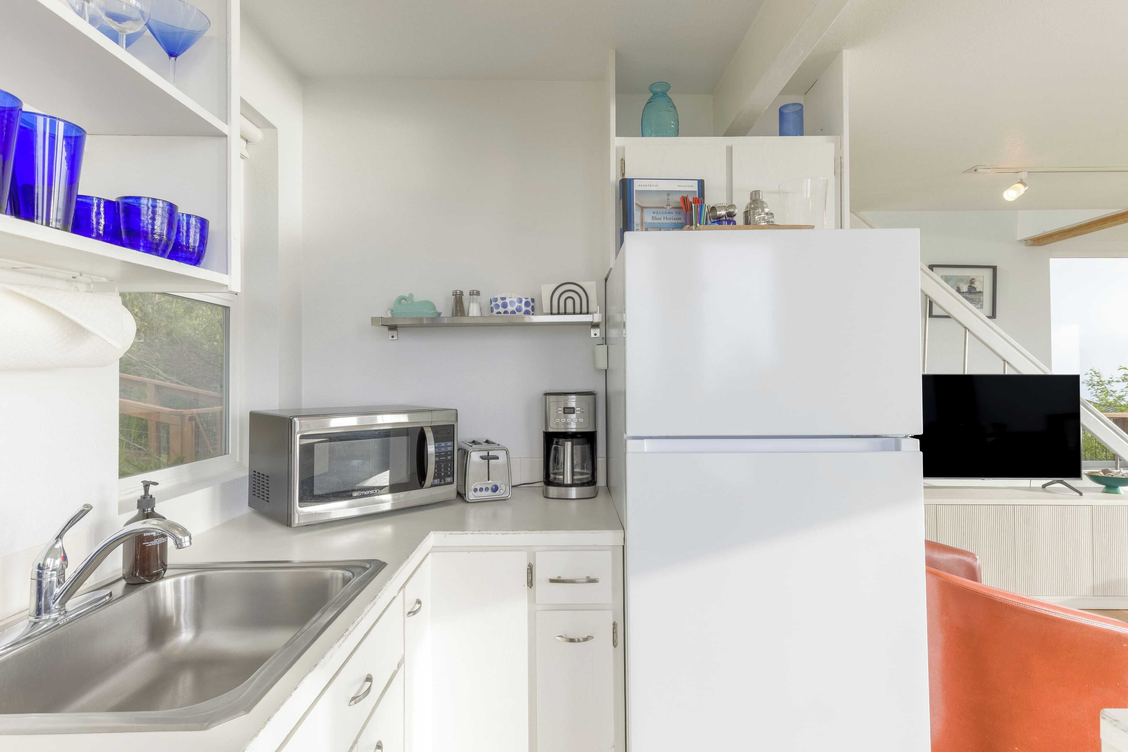 Eine moderne Küche mit weißen Schränken, einer Spüle aus Edelstahl, einer Mikrowelle, einer Kaffeemaschine und einem Kühlschrank. Auf einem Regal stehen blaue Glasvasen, und ein Fenster bietet Blick auf Grünflächen.
