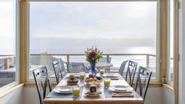 A dining table with breakfast items and a vase of flowers, overlooking a scenic ocean view. - Blue Horizon by Avantstay Panoramic Views of the Ocean w/ Entertainers Patio (Rockaway Beach)