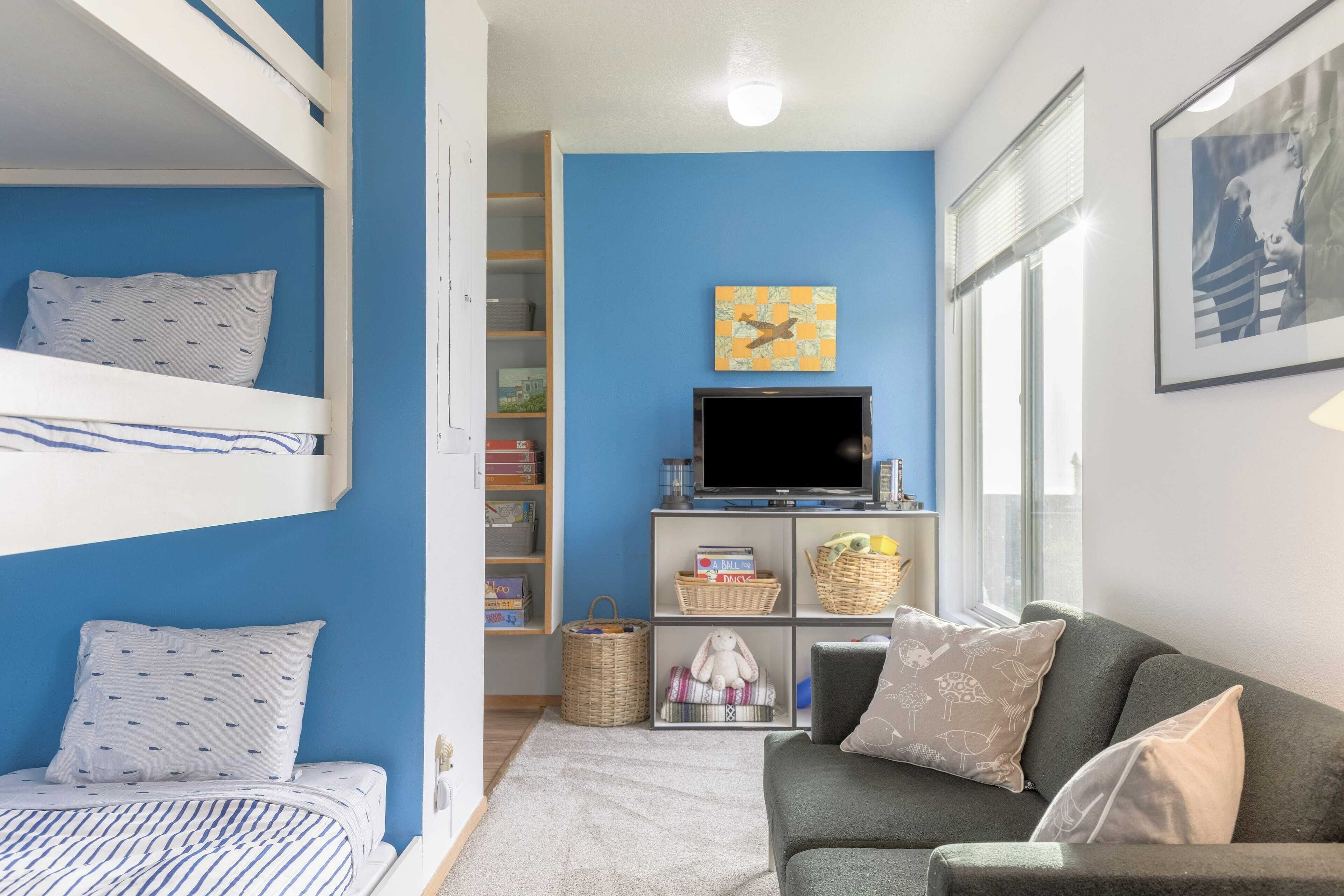 A room with bunk beds, a grey sofa, a TV, and a bookshelf.