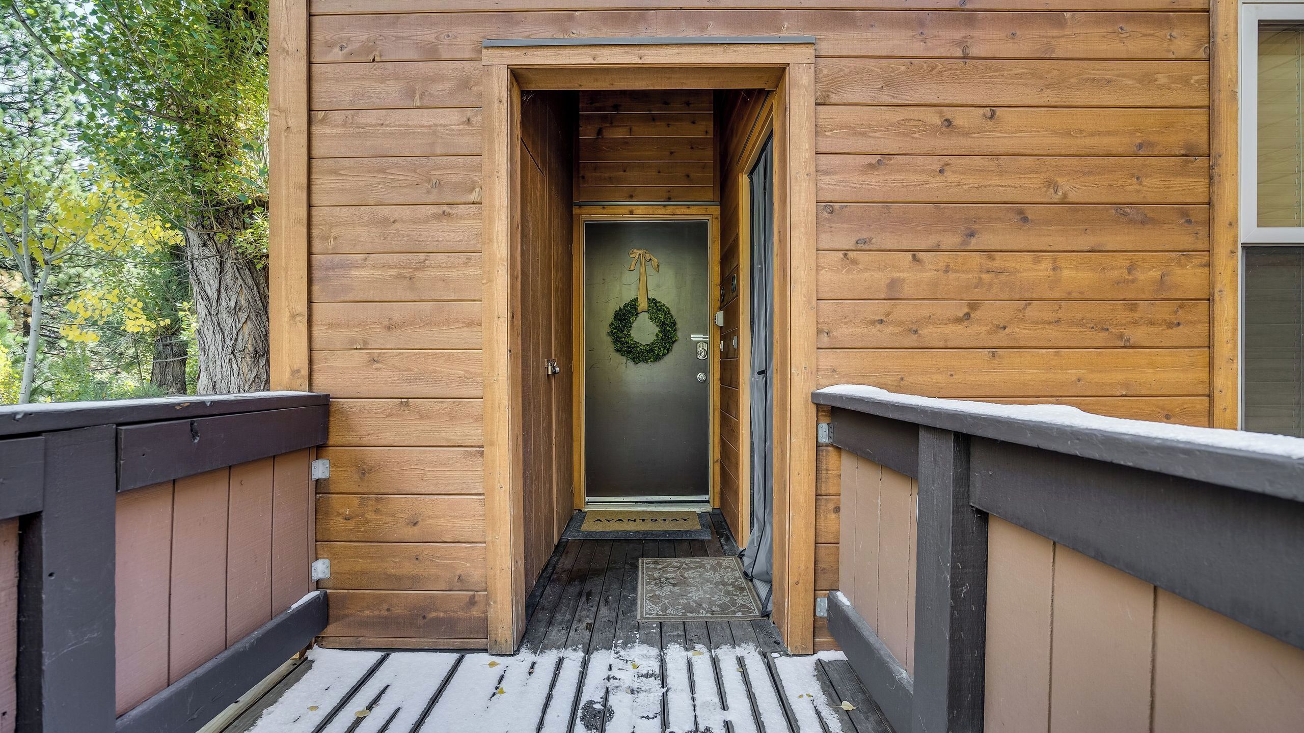 A wooden cabin with a wooden deck, a door with a wreath, and a snow-covered ground.