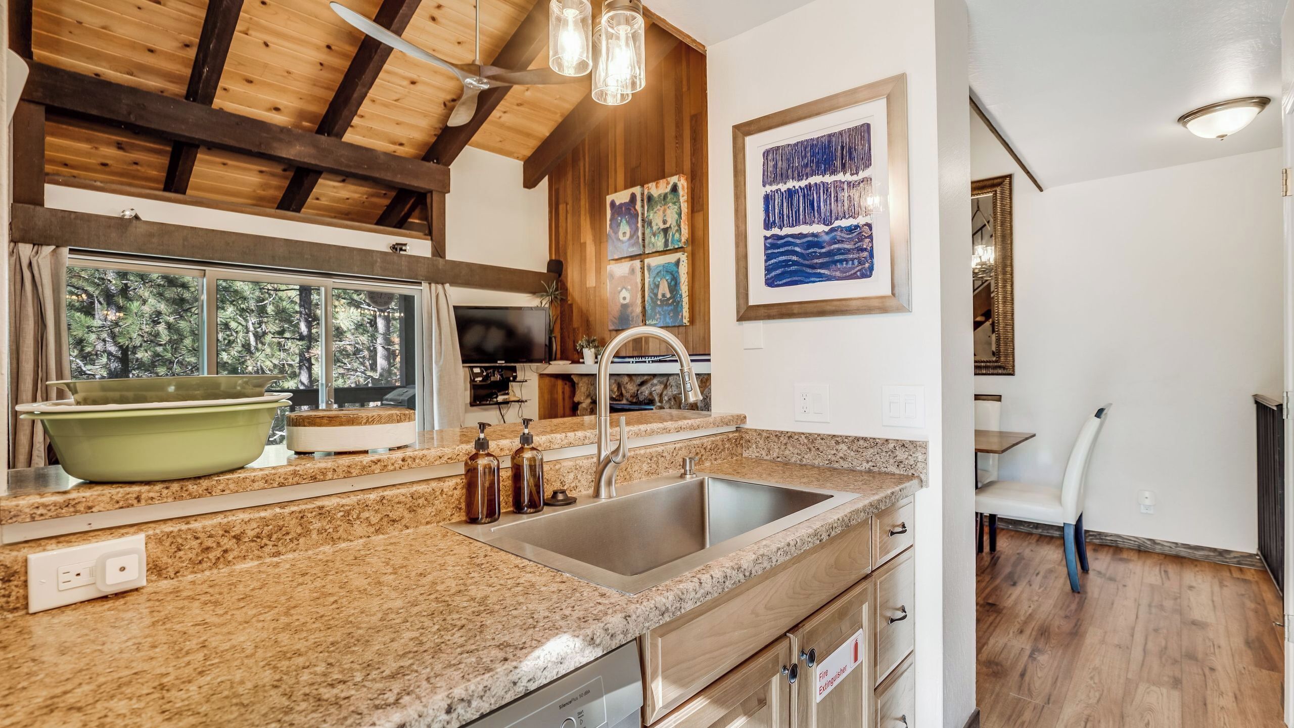 Una cocina con encimeras de granito, pileta de acero inoxidable, vigas de madera en el techo y un ventanal con vista a árboles.