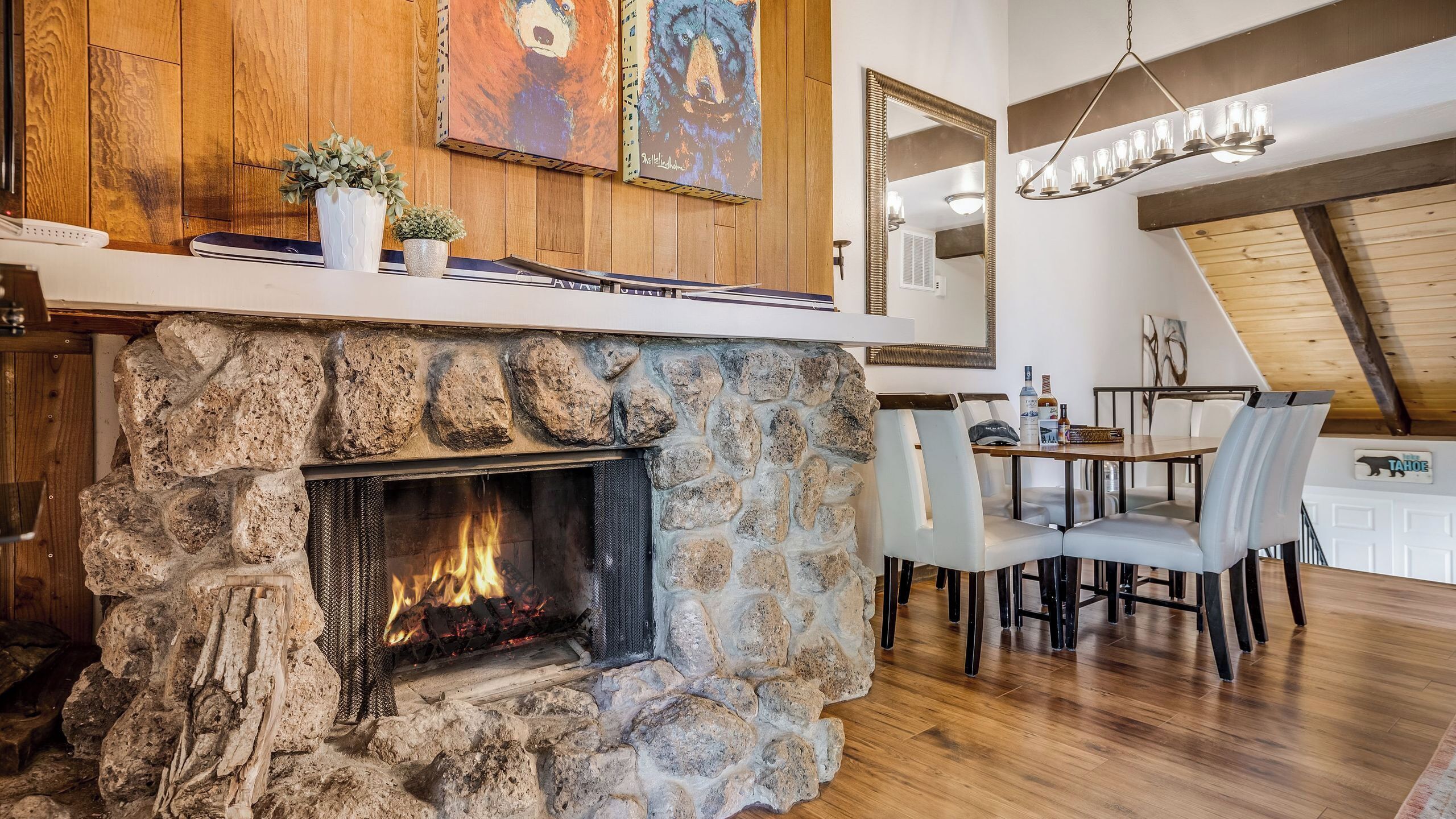 A stone fireplace with a fire, wooden panel walls, a dining area with chairs, and a chandelier.