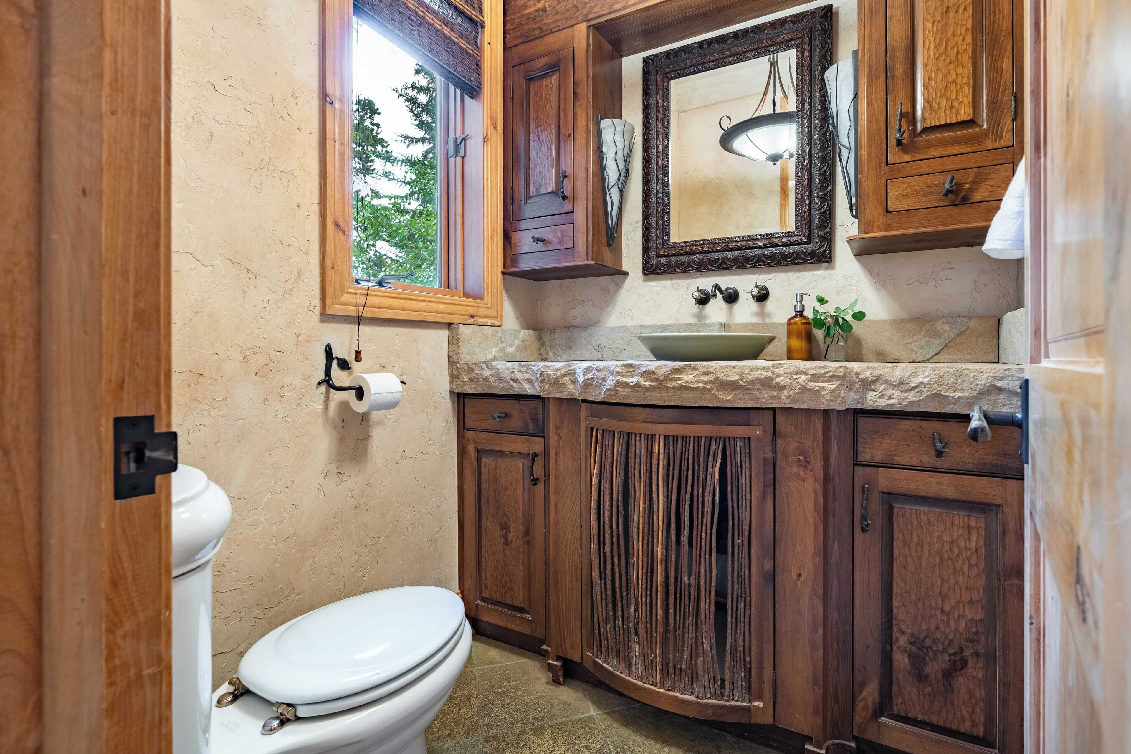 A bathroom with wooden cabinets, a white toilet, a sink with a mirror above it, and a window providing natural light.