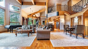 A spacious living area with a grand piano, a fireplace, and a dining area with a high wooden ceiling and a balcony.