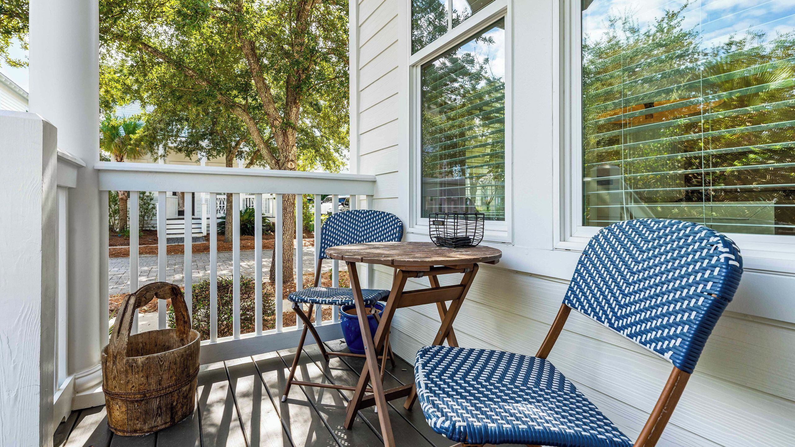 A porch with a wooden table and two blue patterned chairs, a wicker basket, and a window with blinds.