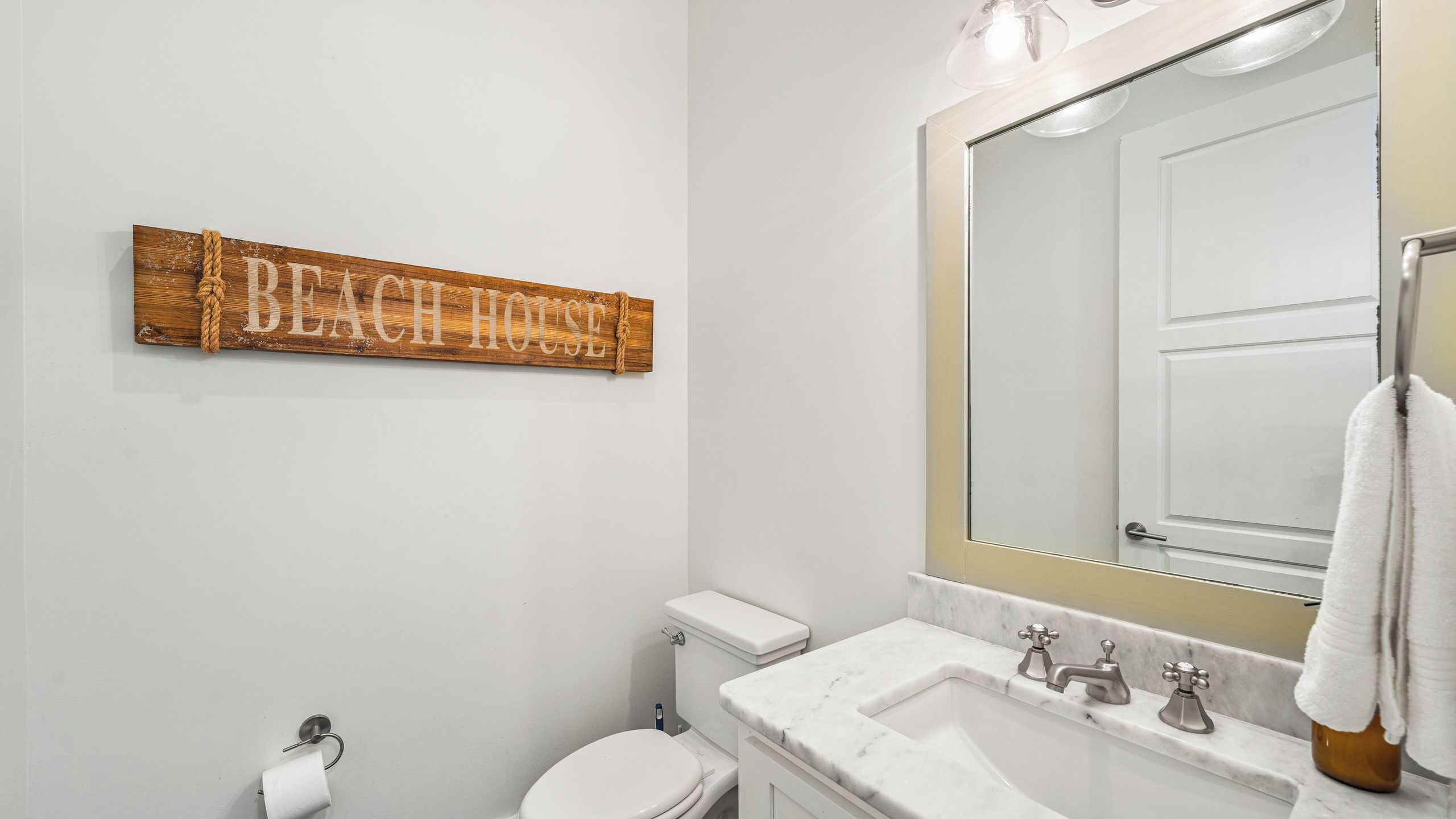 A bathroom with a toilet, sink, and a wooden sign that reads 'BEACH HOUSE'.