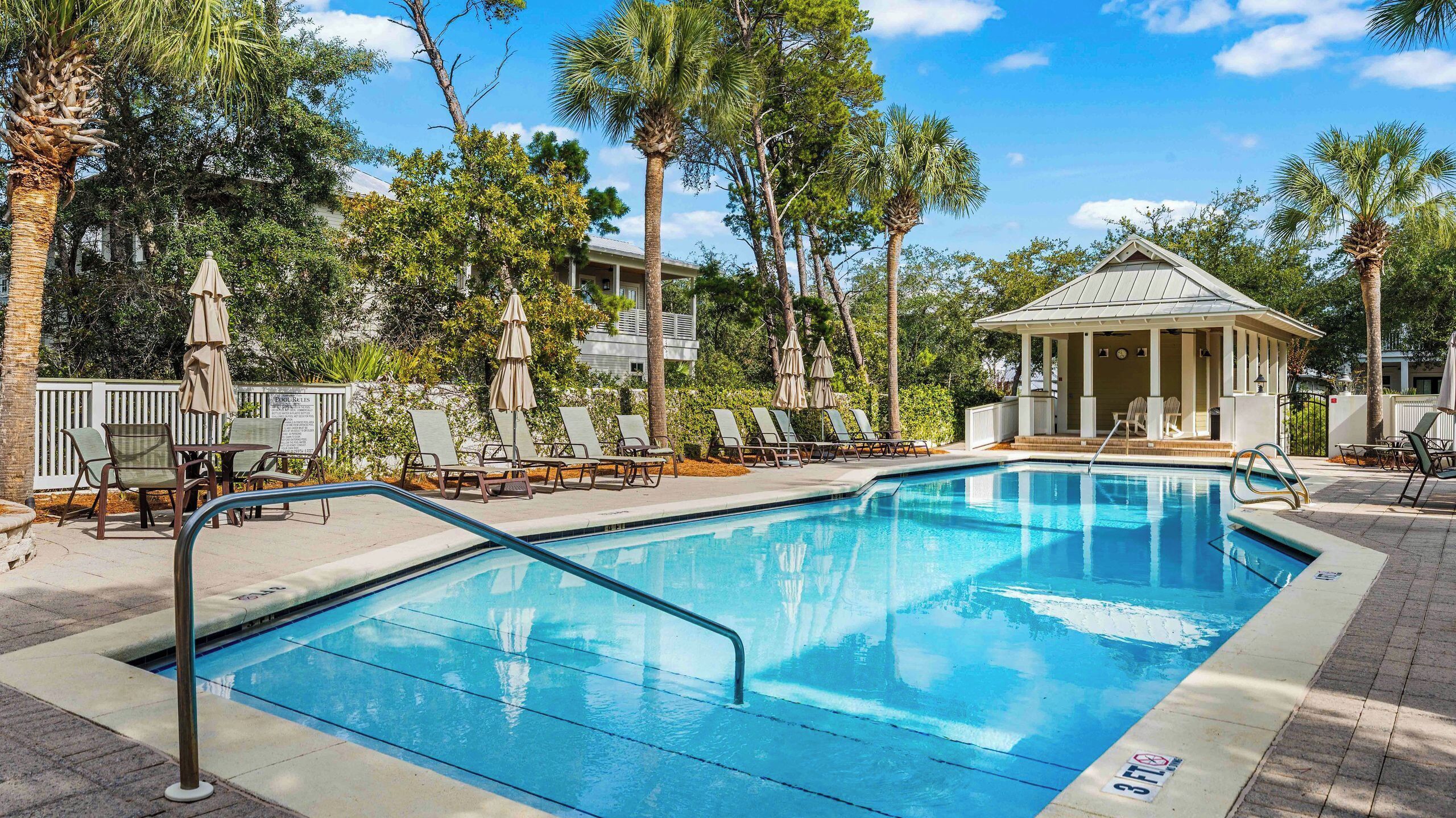 A swimming pool with lounge chairs and umbrellas, surrounded by palm trees and a covered pavilion.