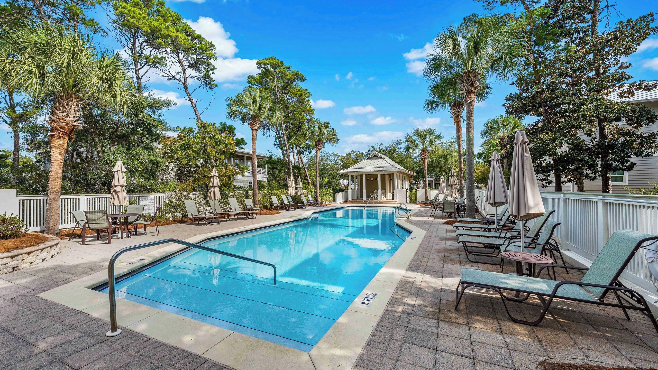 A swimming pool with lounge chairs and umbrellas, surrounded by trees and a building with a covered patio.