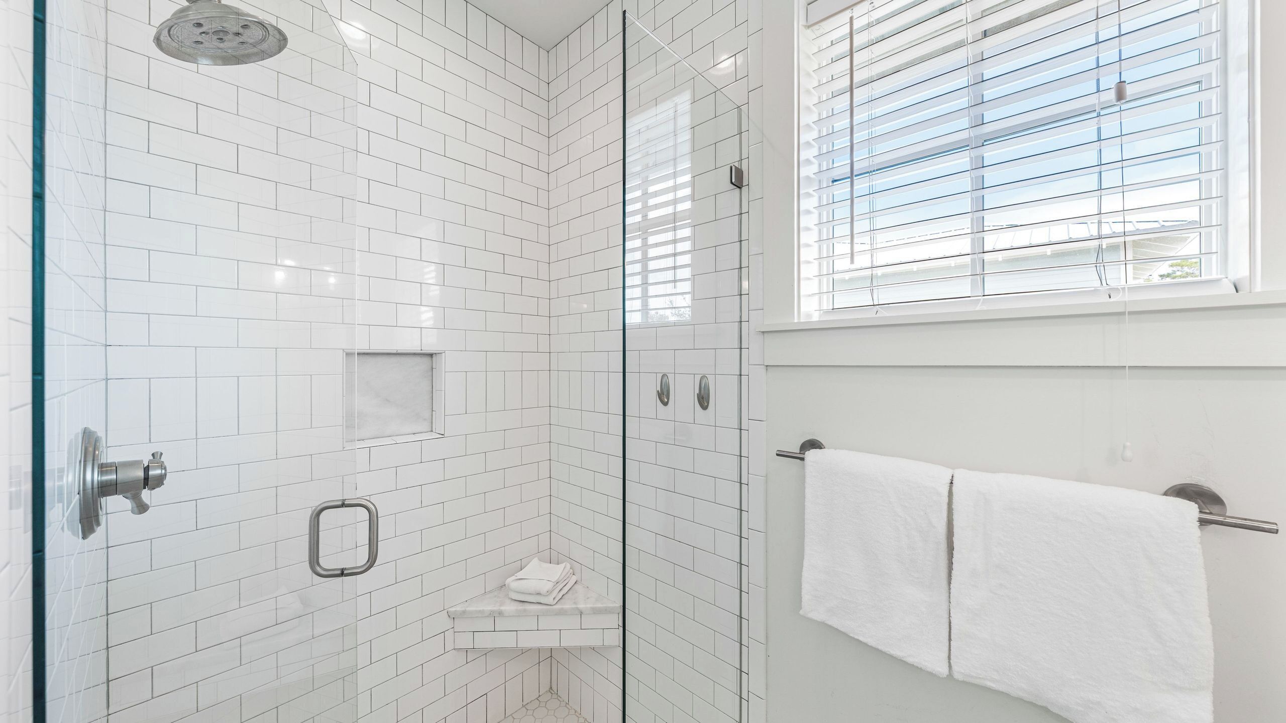 A modern shower with white tiles, a glass door, and a window with blinds.