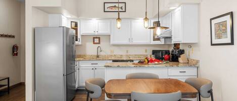 A modern kitchen with white cabinets, a stainless steel refrigerator, a wooden dining table with four chairs, and pendant lighting.