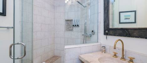 A bathroom with a glass shower enclosure, a sink with a marble countertop, and a mirror with a picture frame.