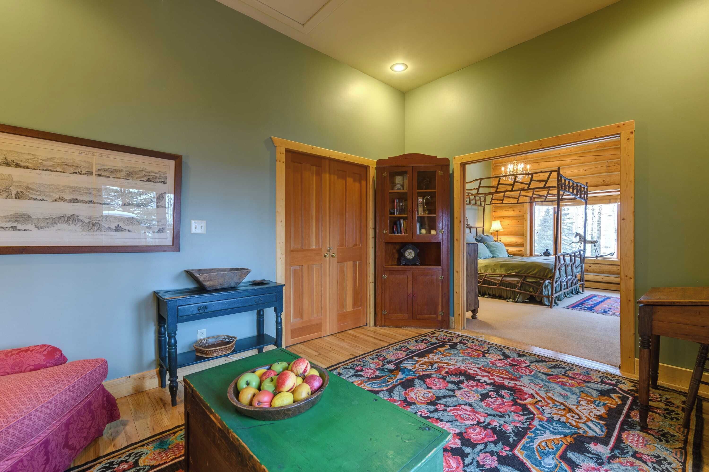 A room with a wooden floor, a green table with a bowl of fruit, a patterned rug, a bed with a wooden ladder, and a wooden cabinet with glass doors.