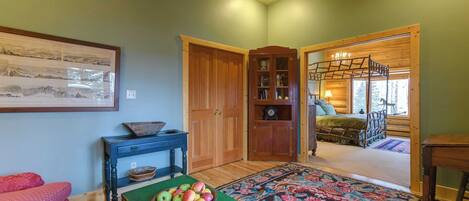 A room with a wooden floor, a green table with a bowl of fruit, a patterned rug, a bed with a wooden ladder, and a wooden cabinet with glass doors.
