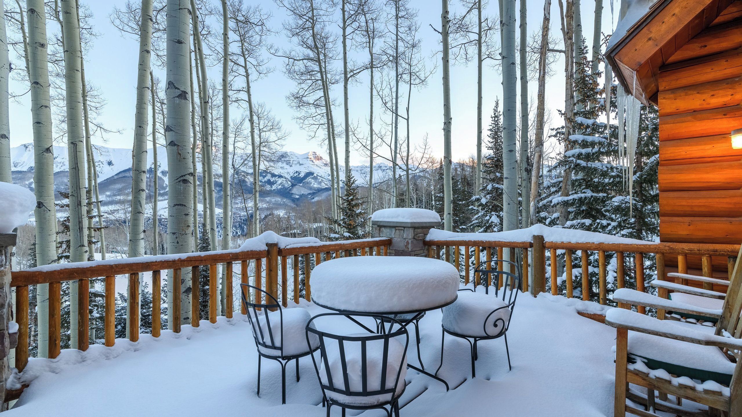 A wooden deck with snow-covered chairs and a table, overlooking a forest and mountains.