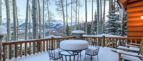 A wooden deck with snow-covered chairs and a table, overlooking a forest and mountains.