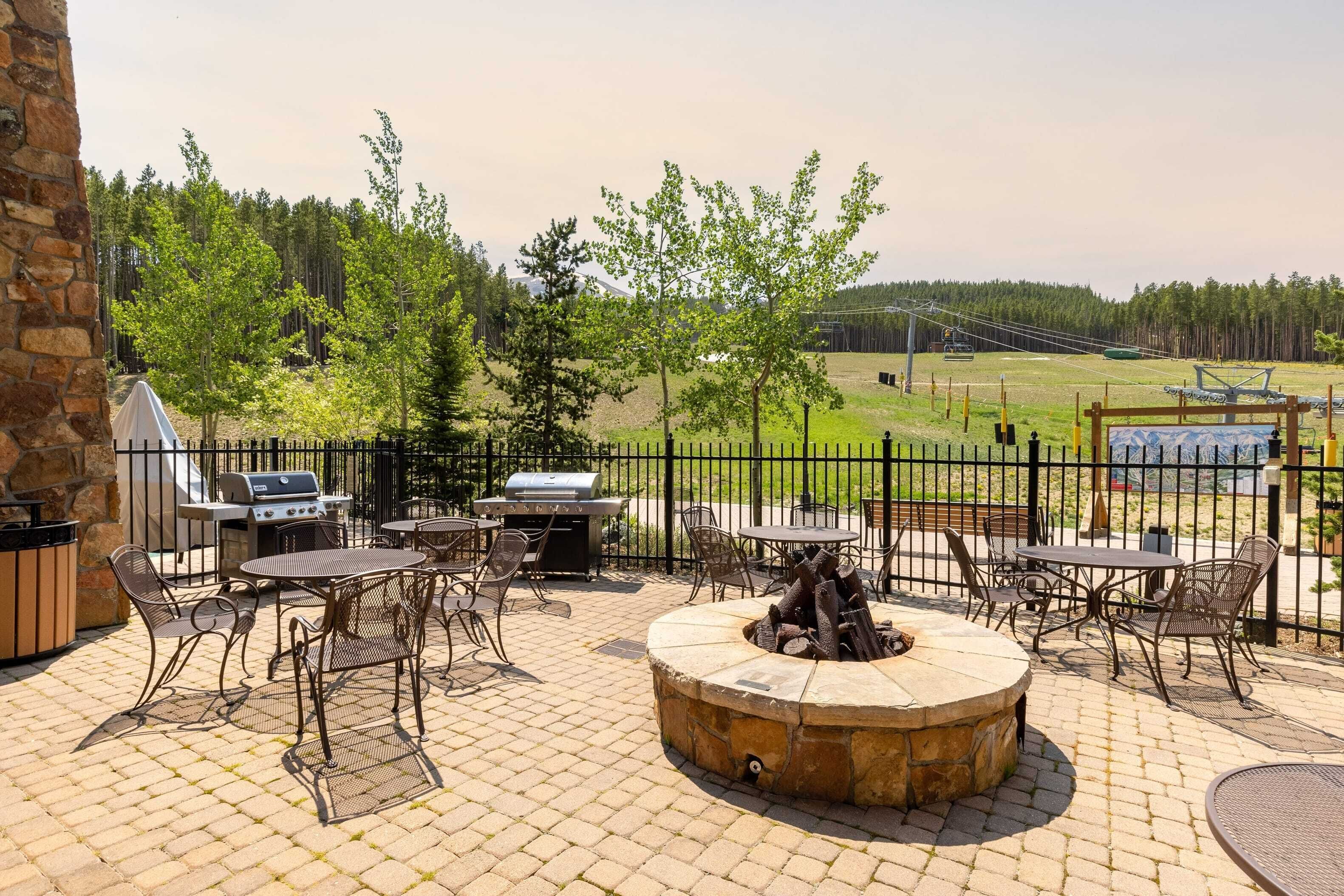 An outdoor patio with a stone fire pit, metal chairs, and a grill area.