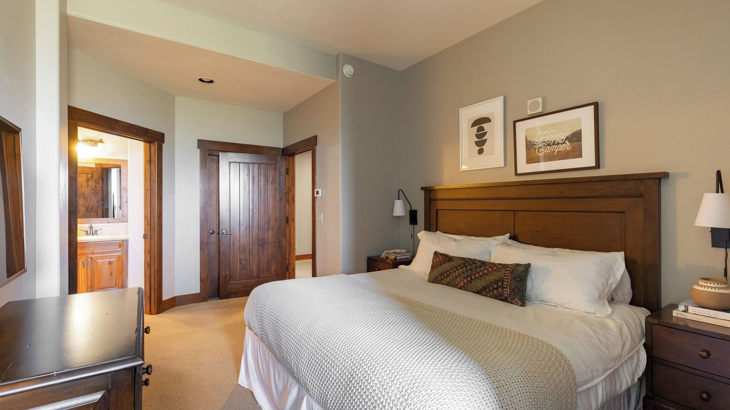 A bedroom with a large bed, wooden headboard, bedside tables, a nightstand, a mirror, and a door leading to another room.