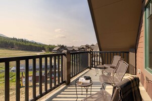 A balcony with outdoor furniture, a view of a forest and mountains, and a building with a green window frame.
