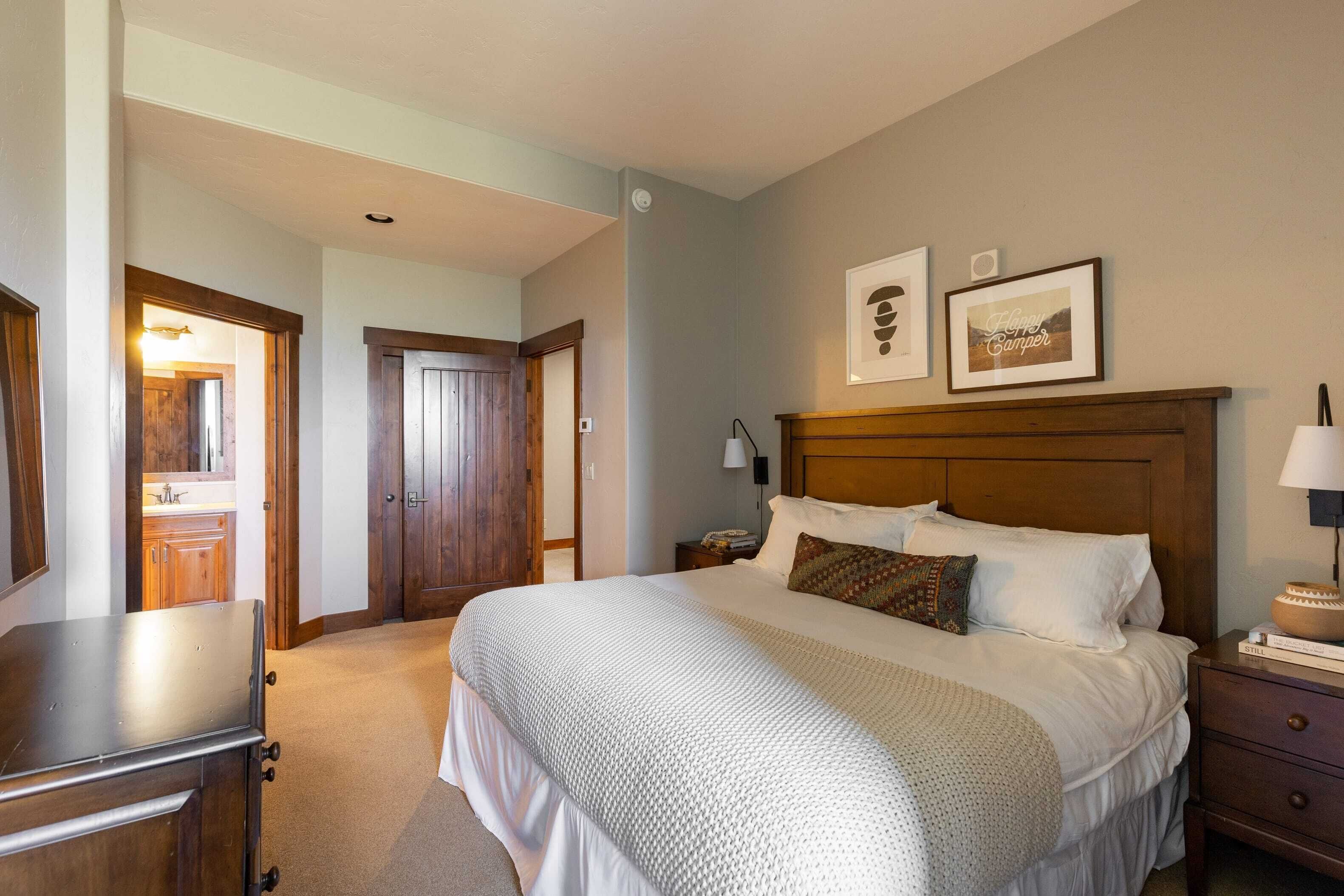 A bedroom with a large bed, wooden headboard, bedside tables, and a nightstand. There is a door leading to another room and a mirror on the wall.