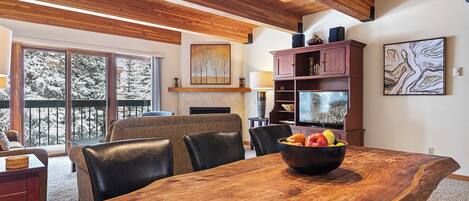 A dining area with a large wooden table, black leather chairs, and a bowl of fruit. There is a fireplace, a television, and a bookshelf in the background.