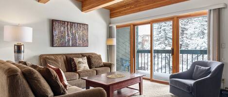 A living room with a wooden ceiling, a large window with a view of snow-covered trees, a brown sofa, a blue armchair, and a wooden coffee table.