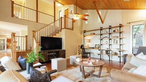 A spacious living room with a wooden staircase, a flat-screen TV, a bookshelf, and a large window. - Dawson by Avantstay Serene Austin Home set Amongst Nature w/ Pool & Close to Lake Travis (Austin)
