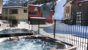 A hot tub with a metal fence in the foreground, a snow-covered street, and colorful buildings in the background. - Cimarron Lodge 20 by Avantstay Ski In/ Ski Out Condo in Ideally Located Complex w/ Hot Tubs! (Telluride)