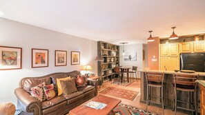 A living room with a leather sofa, a coffee table, and a bookshelf. There is a dining area with a table and chairs. The kitchen has wooden cabinets and a refrigerator.