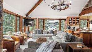 A cozy living room with a fireplace, a television, and a kitchen area with wooden cabinets and a large window.