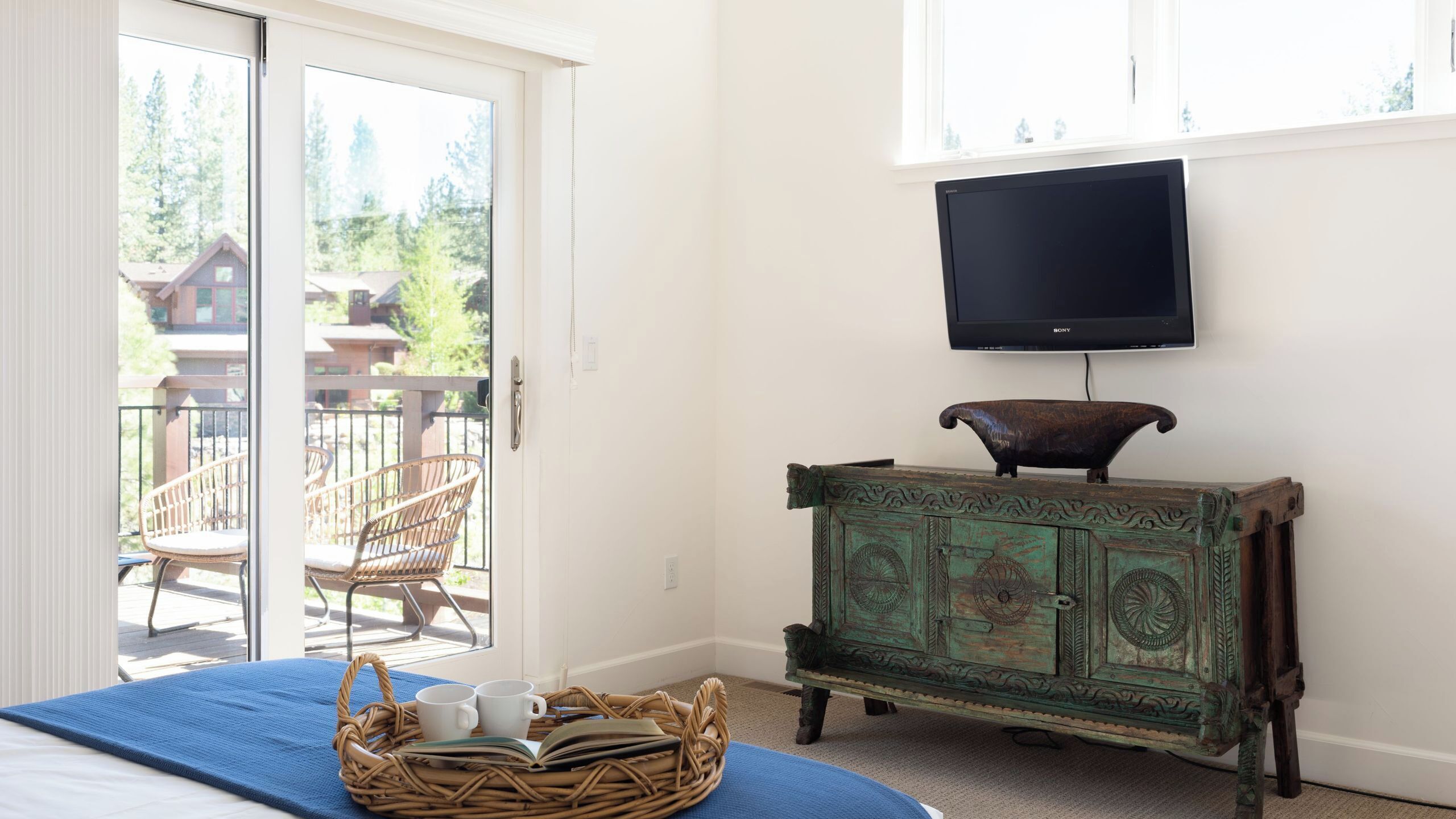 A room with a bed, a wicker basket with cups and a book, a green cabinet with a decorative item, and a TV mounted on the wall.