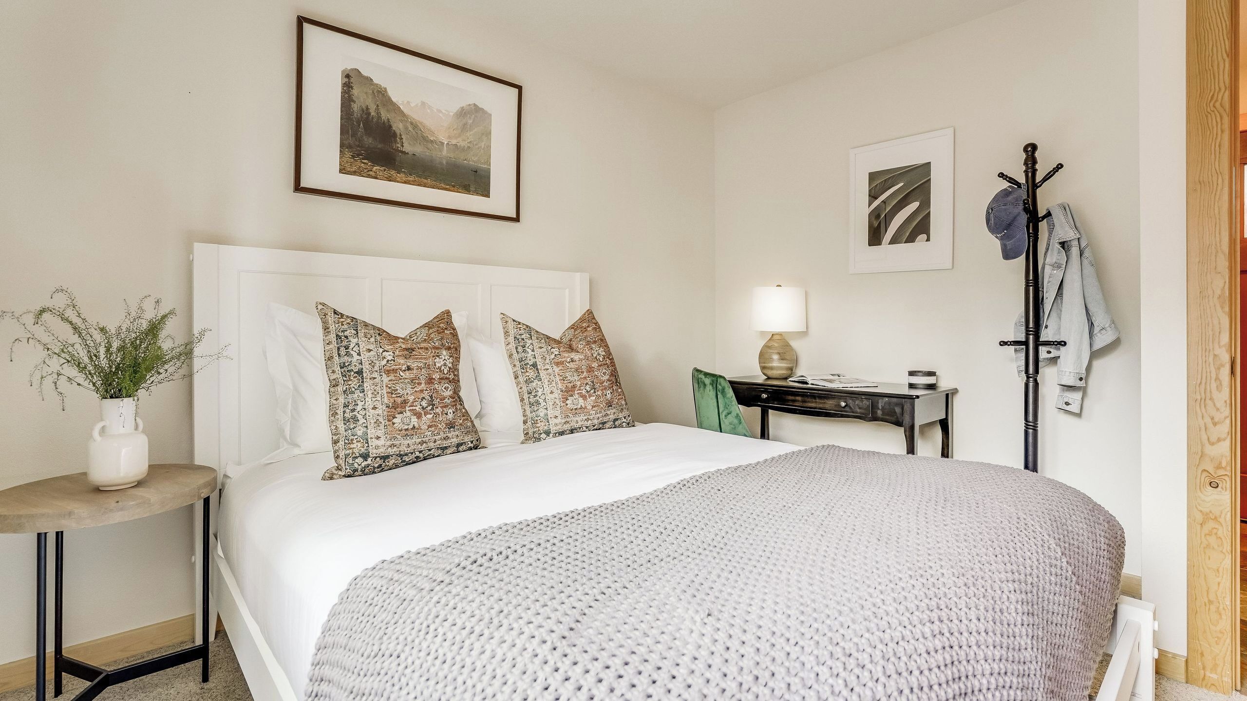 A neatly arranged bedroom with a bed, a bedside table, a lamp, a coat rack, and a framed picture on the wall.