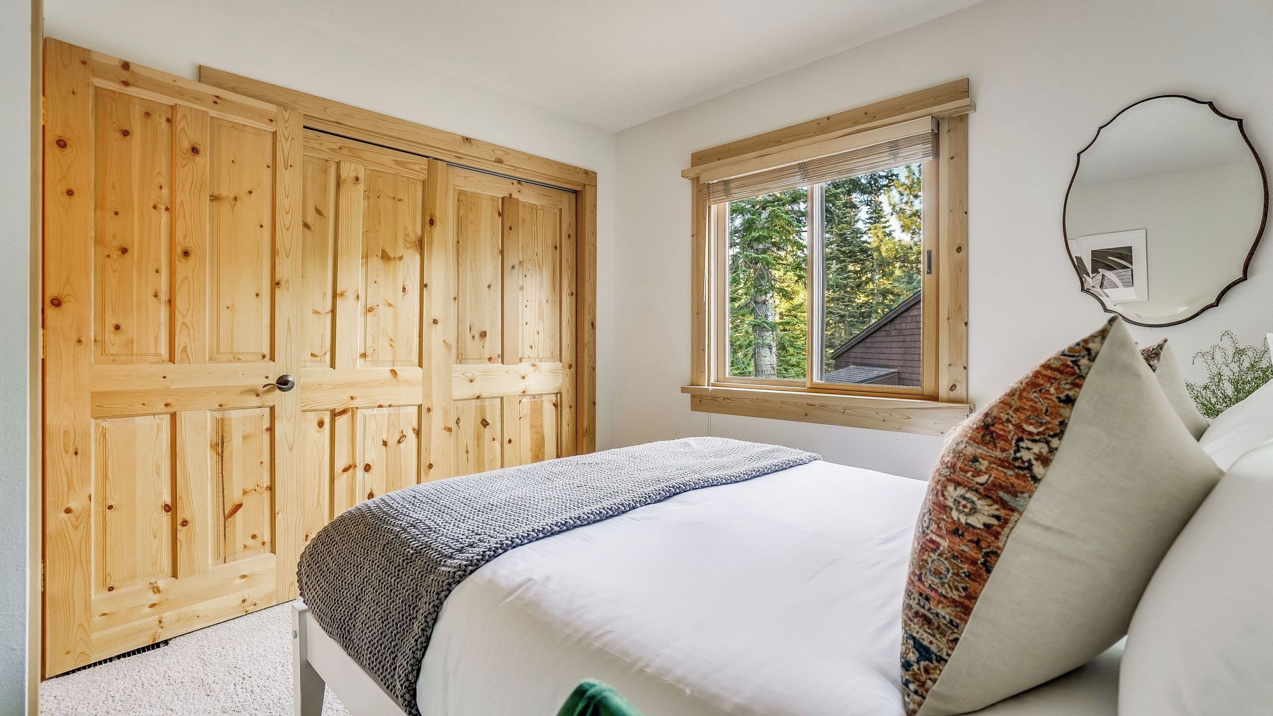 A bedroom with a wooden wardrobe, a window with a view, a bed with pillows, and a mirror on the wall.