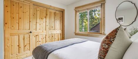 A bedroom with a wooden wardrobe, a window with a view, a bed with pillows, and a mirror on the wall.