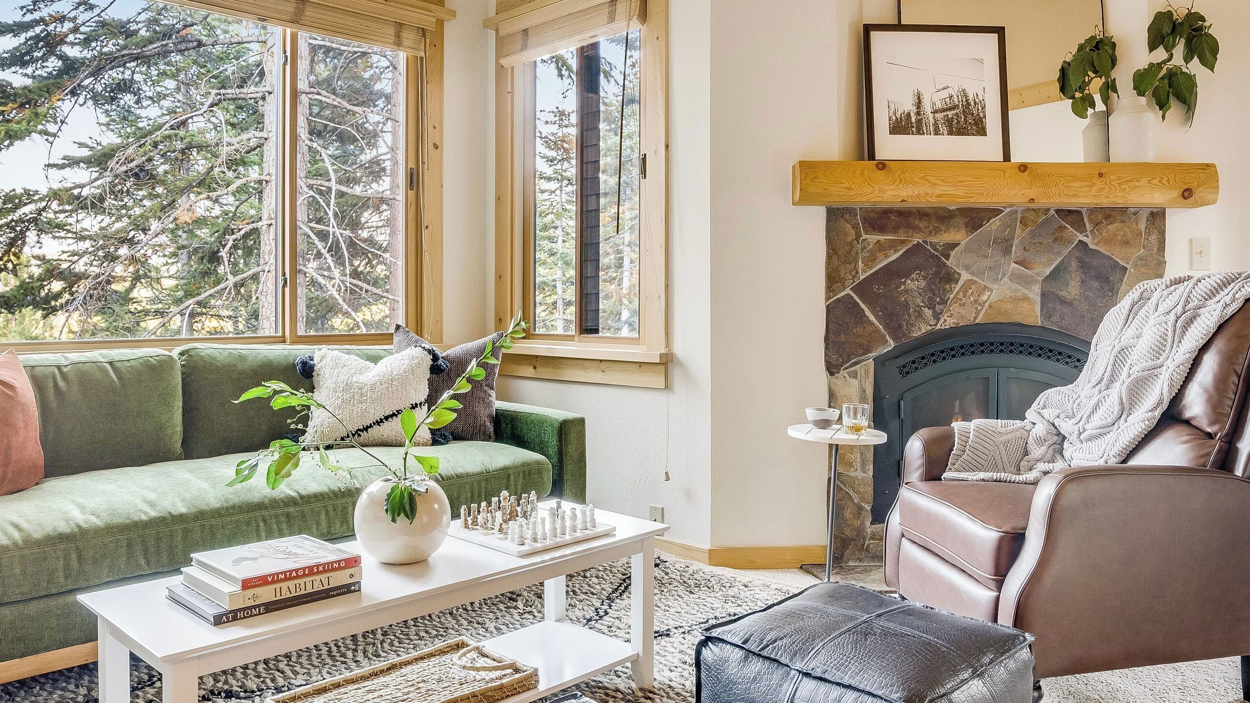 A cozy living room with a green sofa, a white coffee table, a fireplace, and a view of trees through large windows.