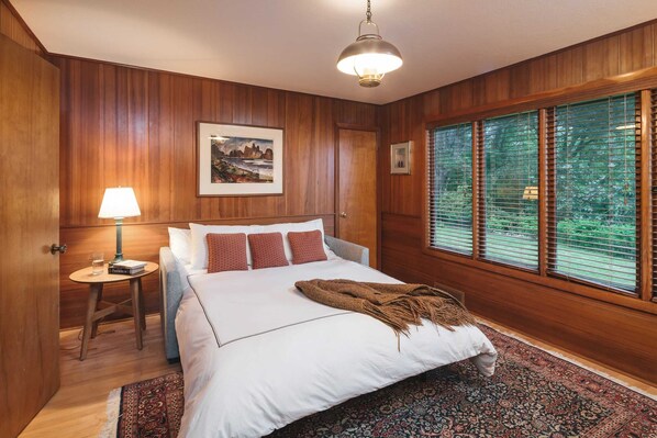 A bedroom with wooden paneling, a bed with white bedding, a lamp, a framed picture, and a window with blinds. - Ocean Views, Outdoor Living, 5min Walk to Beach The Purcell House by Avantstay (Manzanita)