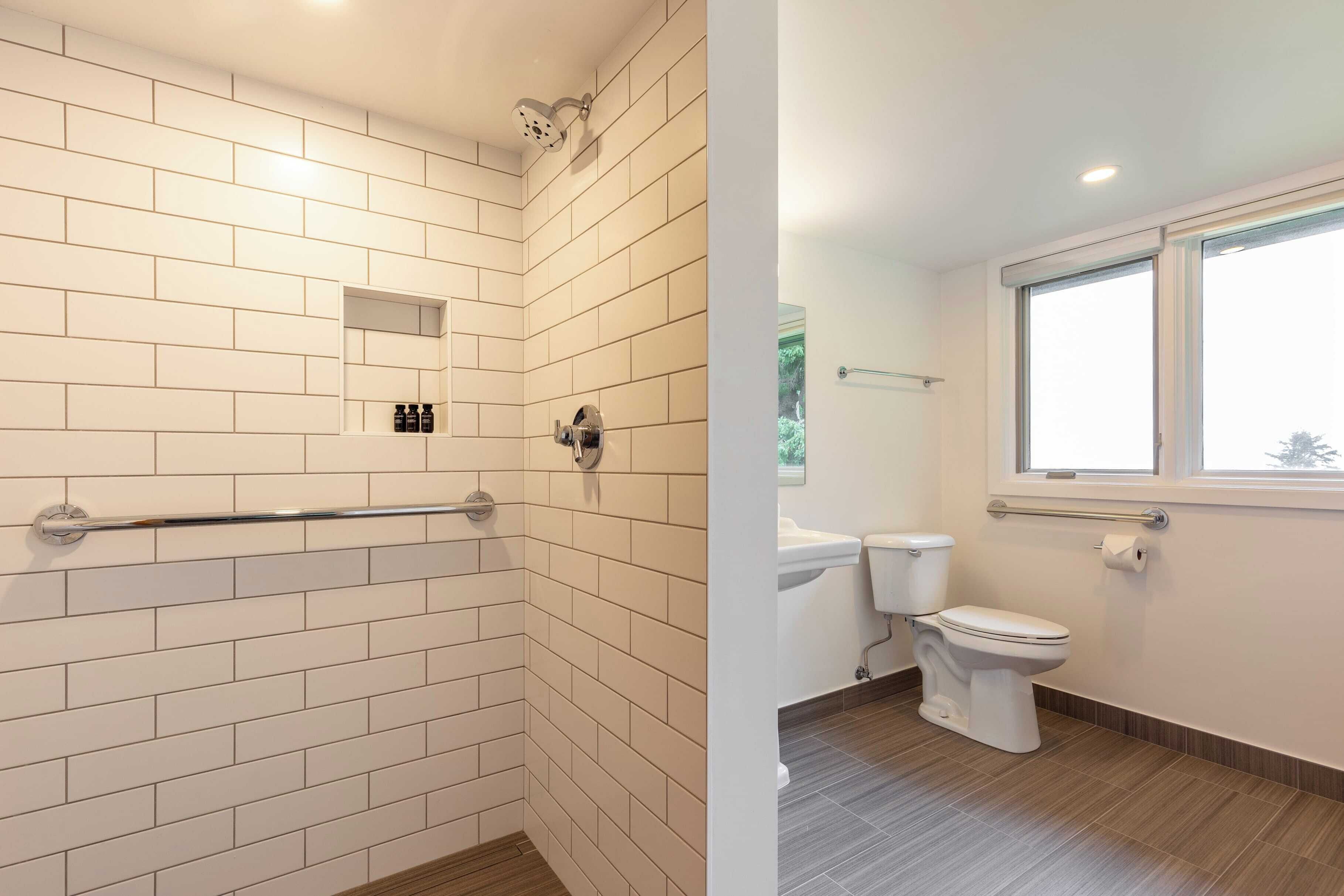 A bathroom with white subway tiles, a shower with a handheld showerhead, a toilet, and a window providing natural light.