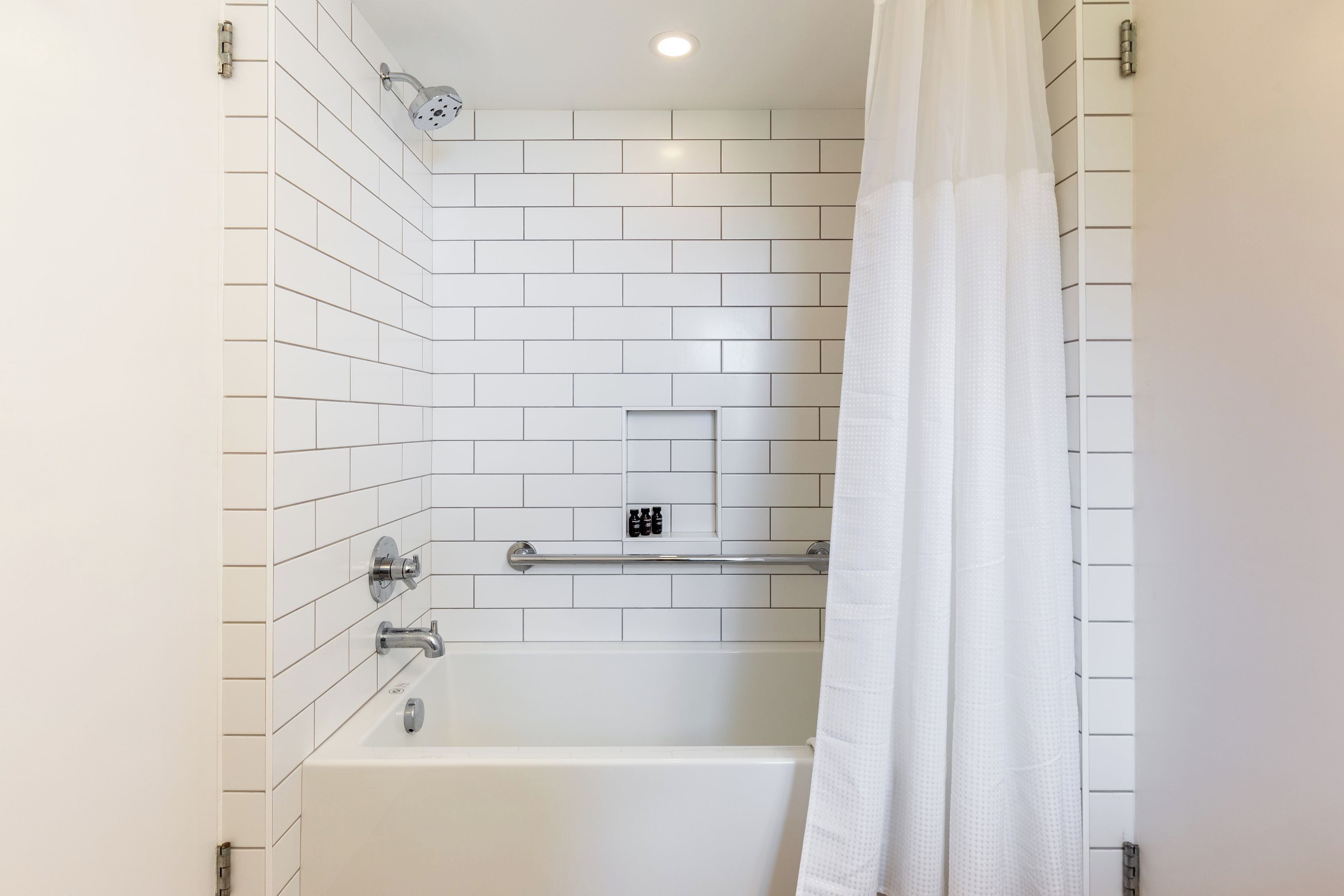 A bathroom with white tiled walls, a bathtub, a showerhead, and a handrail.