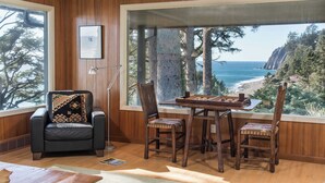 Una habitación con un ventanal grande que da a un paisaje costero, una silla de madera, una mesa con un juego de mesa y un cuadro en la pared.