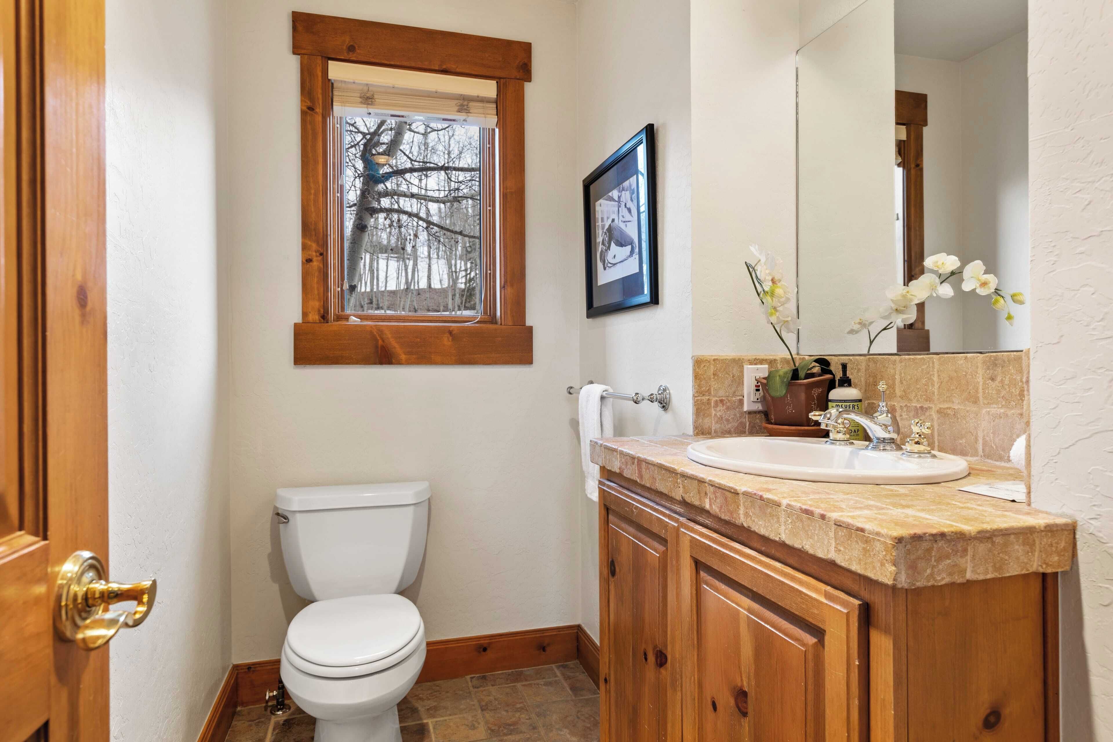 A bathroom with a toilet, wooden vanity, sink, mirror, framed picture, and window.